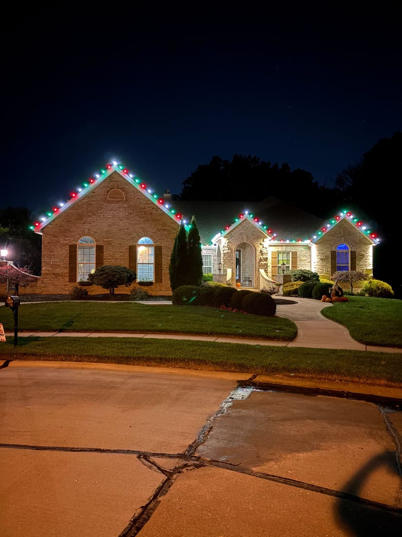 Holiday-decorated brick house at night with colorful lights and landscaped yard.